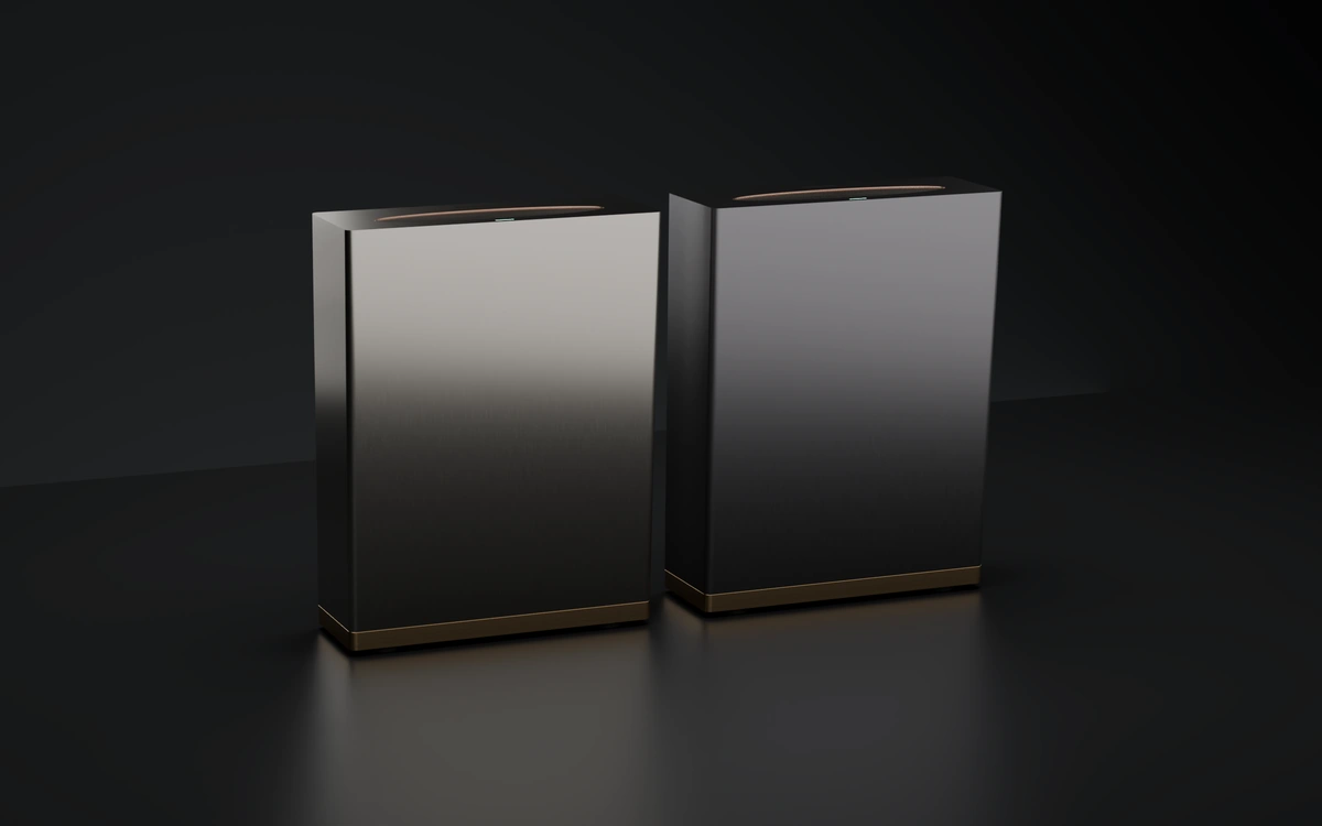 Render of two Ameka One machines side by side: a brushed silver one on the left and a dark gunmetal one on the right, both with bronze bases.